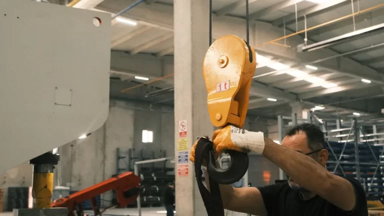 Pracownik w hali produkcyjnej zakłada taśmę nośną na hak suwnicy.